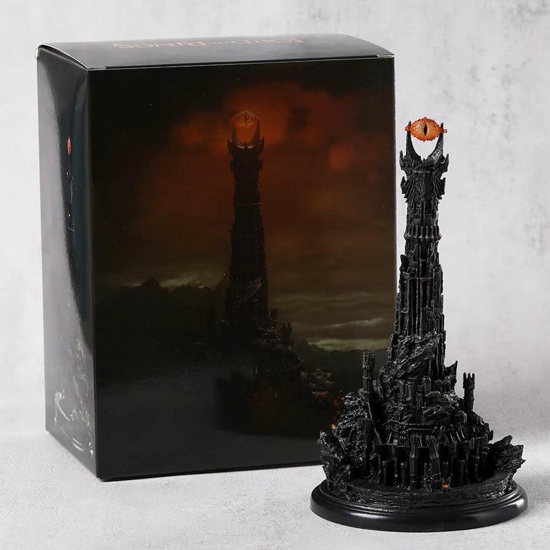 May include: A black sculpture of a tall tower with an orange eye at the top, next to a black box with a similar image. The tower has intricate details and sits on a round base. The box has the words "LORD OF THE RINGS" on top.