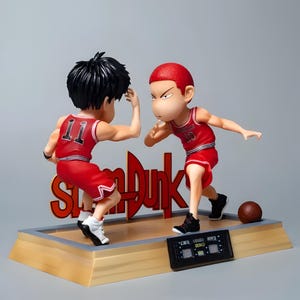 May include: Two red and white basketball player figurines, one with the number 11 and the other with the number 10, are posed in a basketball game action pose. The figures are on a wooden base with a digital scoreboard and the word "Slam Dunk" in red letters.