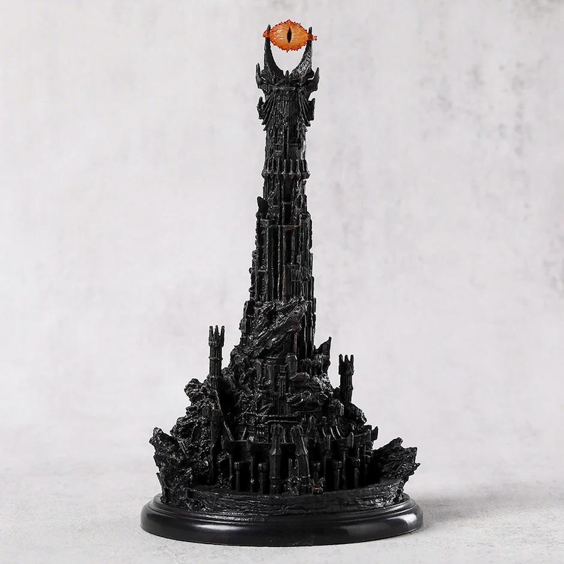 May include: A black, detailed sculpture of a tall tower with an orange eye at the top. The tower has intricate architectural details and sits on a round base. The eye is a prominent feature, adding a striking contrast to the dark structure.