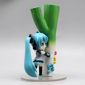 May include: A figurine of a blue-haired anime character with headphones, leaning against a large green and white leek. The figurine has a gray top, black skirt, and black boots. Musical notes are attached to the leek.