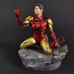 May include: A red and gold Iron Man action figure in a kneeling pose, with a damaged suit and a distressed base. The figure is made of plastic and has a detailed paint job.