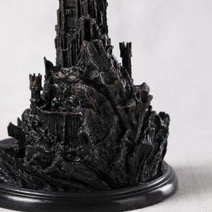 May include: A detailed, dark-colored miniature castle sculpture. The castle features intricate architectural details, including towers, walls, and a rocky base. The sculpture is mounted on a round, black base, and the overall aesthetic is gothic and imposing.