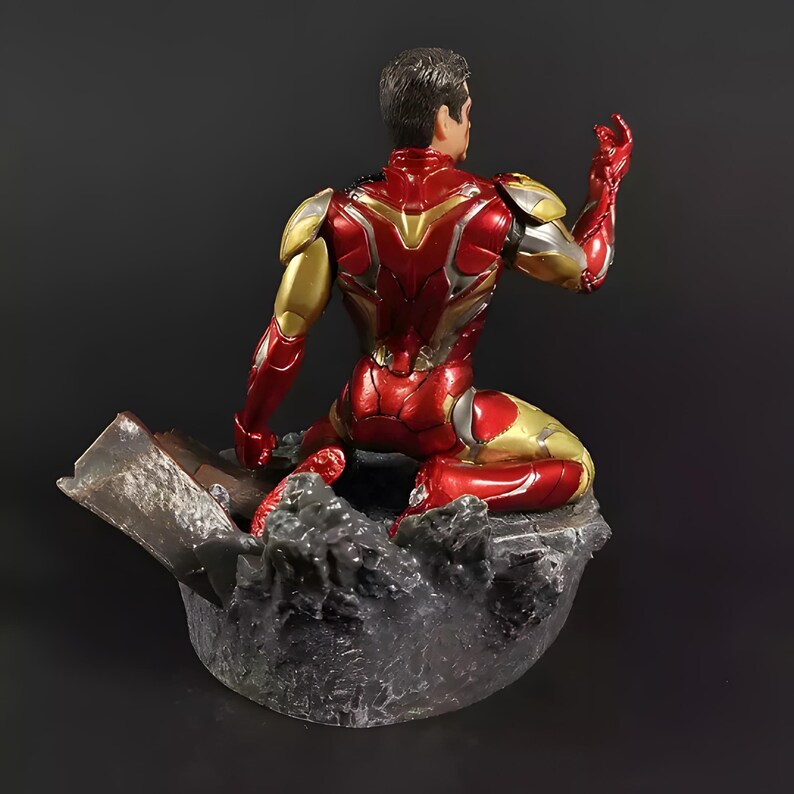 May include: A red and gold Iron Man action figure in a seated pose, with a detailed metallic finish. The figure is posed on a rocky base, with a piece of debris behind it.