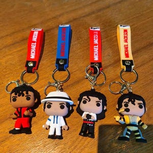 May include: Four keychains featuring pop figures of Michael Jackson in various iconic outfits. Each keychain has a colored strap with "MICHAEL JACKSON" printed on it. The figures are attached to a metal ring.