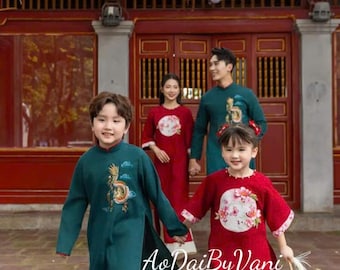 May include: A family of four wearing traditional Vietnamese clothing, called Ao Dai, in red and green colors with embroidered designs. The family is standing in front of a red building with a green roof.