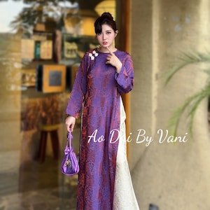 May include: A long, purple dress with a floral pattern and long sleeves. The dress has a white lace panel on the side. A small, purple drawstring bag is held in the hand. The text "Ao Dai By Vani" is visible.