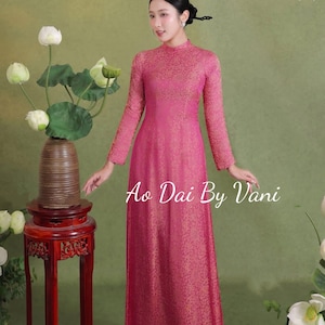 May include: A woman wearing a long-sleeved, magenta-colored Ao Dai dress with a high neckline. The dress has a subtle floral pattern. The text "Ao Dai By Vani" is visible on the dress. The background is a muted green with floral accents.