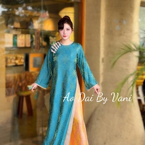 May include: A teal Ao Dai dress with long sleeves and a floral pattern. The dress has a high slit and is paired with orange and yellow trousers. The text "Ao Dai By Vani" is visible on the dress.