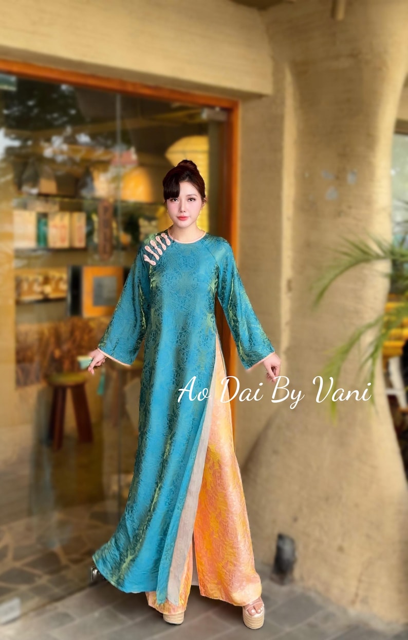 May include: A teal Ao Dai dress with long sleeves and a floral pattern. The dress has a high slit and is paired with orange and yellow trousers. The text "Ao Dai By Vani" is visible on the dress.