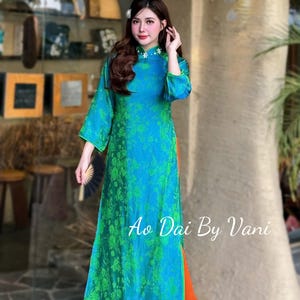 May include: A long, flowing teal and green floral print dress with orange pants. The dress has a high collar with white floral accents and long sleeves. The text "Ao Dai By Vani" is visible on the lower right of the dress.
