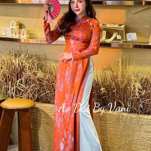 May include: A woman wearing a long, orange dress with a floral pattern and a light blue underlay. She holds a pink fan. The dress has long sleeves and a mandarin collar. The text "Ao Dai By Vani" is visible.
