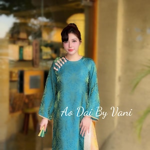 May include: Teal Ao Dai dress with a floral pattern and long sleeves. The dress has a mandarin collar and a side slit. The text "Ao Dai By Vani" is displayed on the right side of the dress.