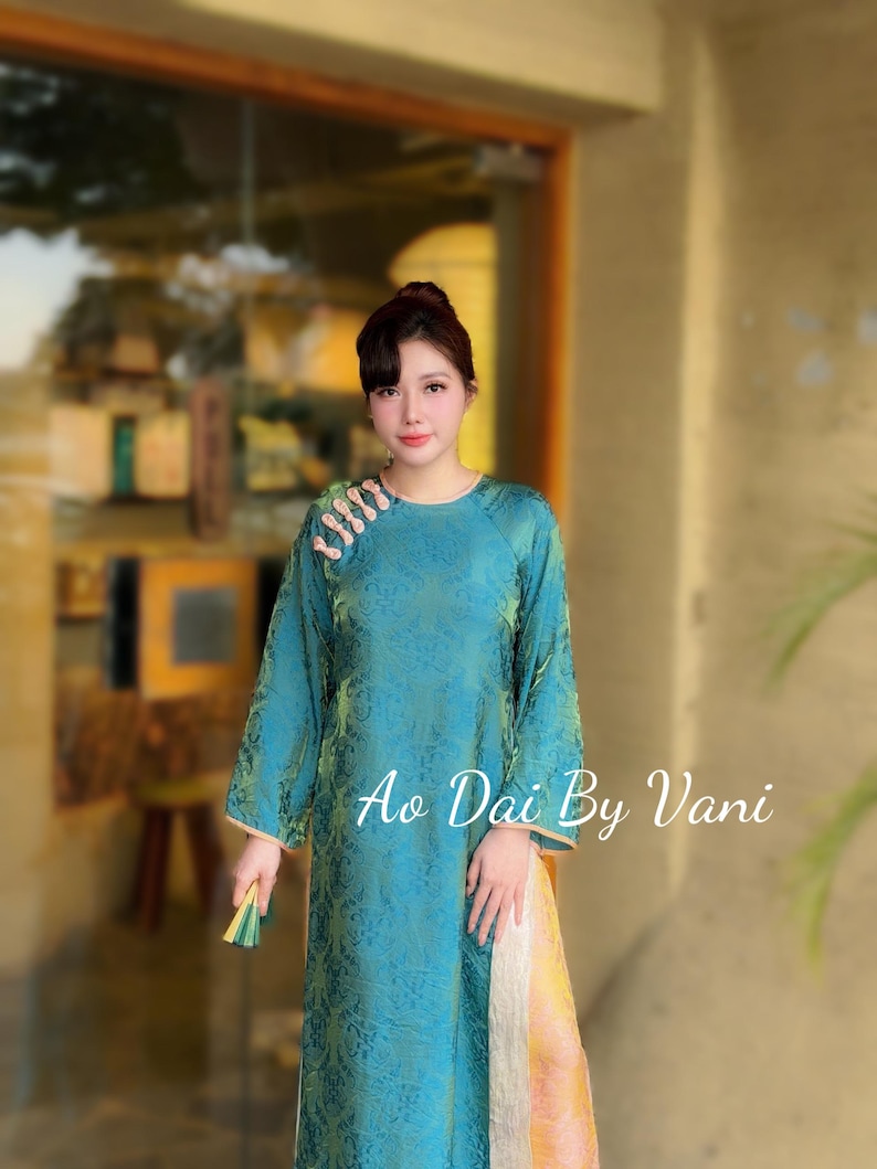 May include: Teal Ao Dai dress with a floral pattern and long sleeves. The dress has a mandarin collar and a side slit. The text "Ao Dai By Vani" is displayed on the right side of the dress.