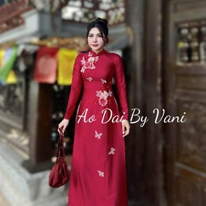 May include: A long, red Ao Dai dress with floral and butterfly accents. The dress has long sleeves and a high collar. The text "Ao Dai By Vani" is displayed on the dress. A matching red handbag is held in the hand.