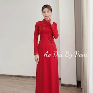 May include: A long, vibrant red dress with long sleeves and a high collar. Floral embellishments adorn the bodice and sleeve. The text "Ao Dai By Vani" is visible on the dress. The dress is floor-length and appears to be made of a smooth fabric.