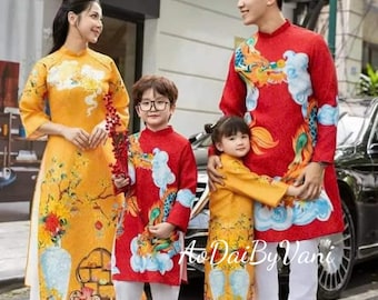 May include: A family of four wearing traditional Vietnamese clothing, known as Ao Dai, in red, yellow, and white colors with dragon and cloud patterns. The clothing features long sleeves and a mandarin collar. The family is standing in front of a building with a sign that says "Ao Dai By Vani".