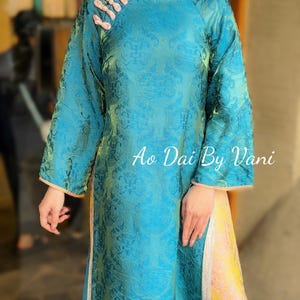 May include: A teal Ao Dai dress with a floral pattern and long sleeves. The dress has a mandarin collar with pink accents and the text "Ao Dai By Vani". The dress has a yellow and pink panel on the side.