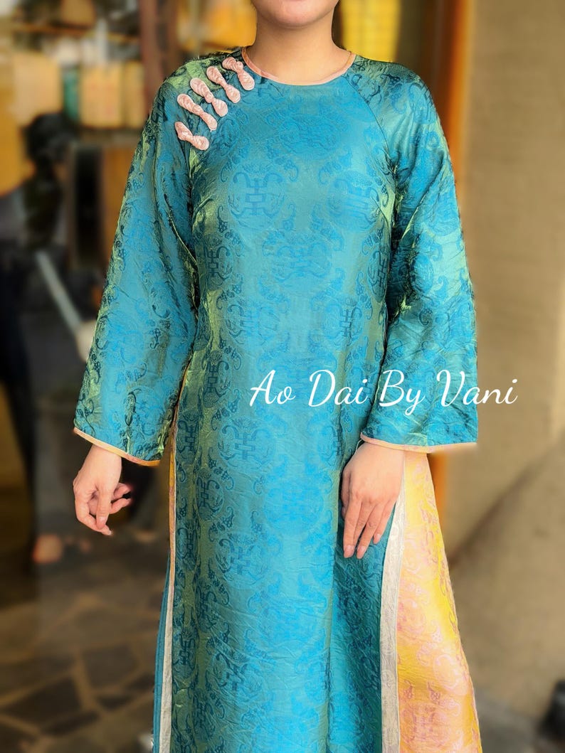 May include: A teal Ao Dai dress with a floral pattern and long sleeves. The dress has a mandarin collar with pink accents and the text "Ao Dai By Vani". The dress has a yellow and pink panel on the side.