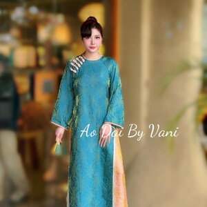 May include: A teal-coloured, long-sleeved dress with a floral pattern and a high neckline. The dress has a side slit and is embellished with decorative details on the shoulder. The text "Ao Dai By Vani" is visible on the dress.