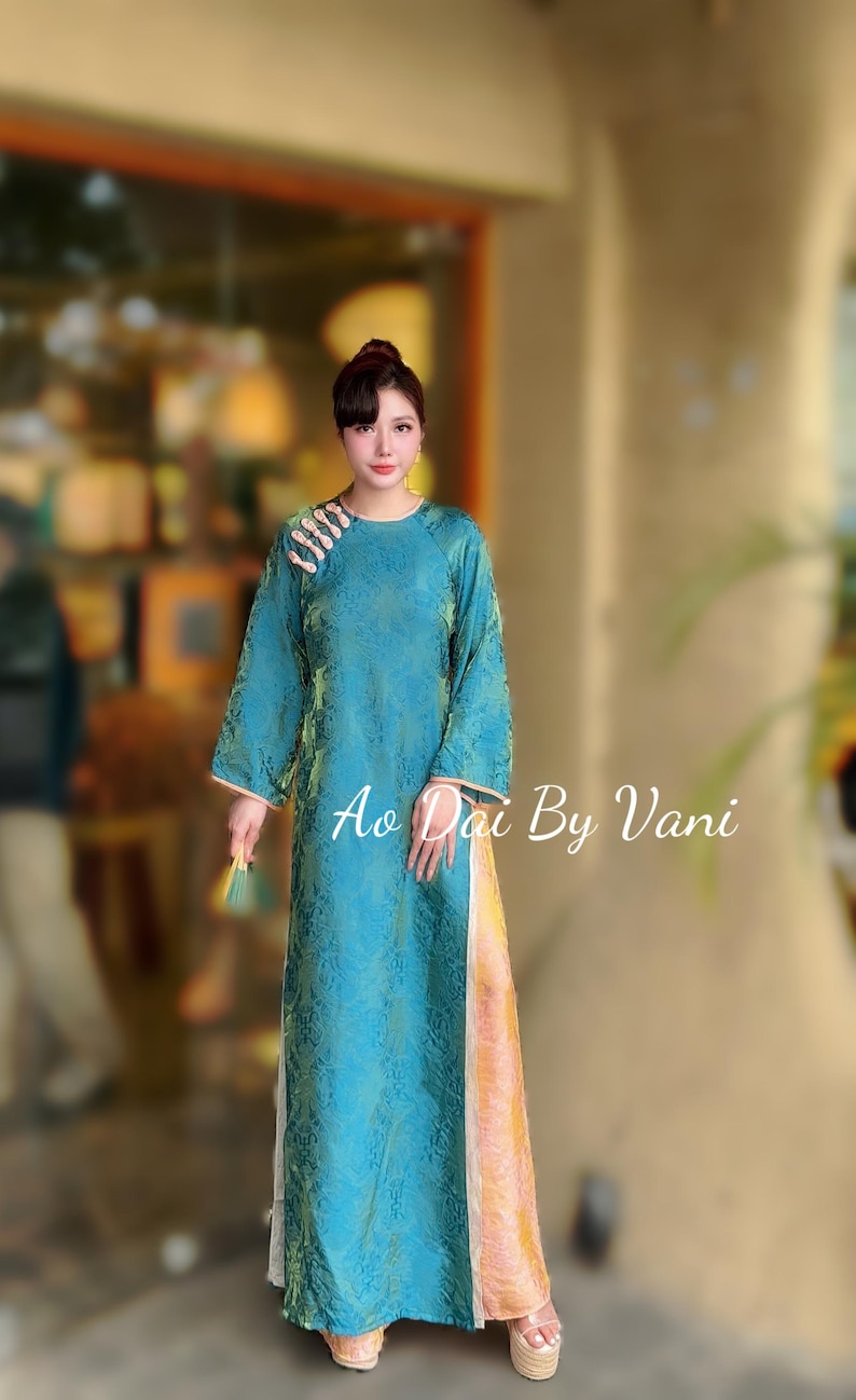 May include: A teal-coloured, long-sleeved dress with a floral pattern and a high neckline. The dress has a side slit and is embellished with decorative details on the shoulder. The text "Ao Dai By Vani" is visible on the dress.