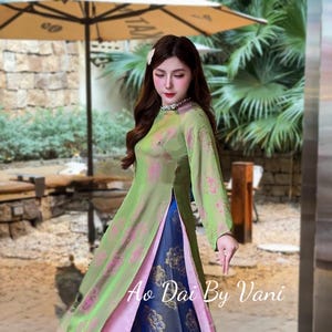May include: A woman wearing a traditional Vietnamese Ao Dai dress. The dress features a light green overcoat with pink floral patterns, a pink underlayer, and blue pants with gold floral designs. The text "Ao Dai By Vani" is visible.