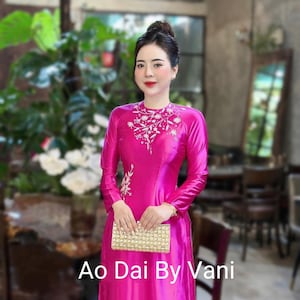 May include: A vibrant pink Ao Dai dress with long sleeves and intricate white embroidery on the neckline and chest. The dress is paired with a gold clutch. The text "Ao Dai By Vani" is visible.