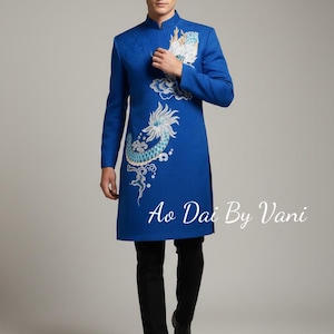 May include: A vibrant blue tunic with a mandarin collar and long sleeves, featuring an embroidered dragon design in white and teal. The tunic is paired with black pants. Text on the image reads "Ao Dai By Vani".
