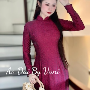 May include: A long-sleeved, burgundy dress with a high collar. The dress has a textured pattern and is paired with a woven handbag adorned with white floral accents. The text "Ao Dai By Vani" is visible.