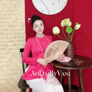 May include: A woman wearing a pink traditional Vietnamese dress, called an áo dài, with white pants. She is holding a wooden fan and sitting on a wooden chair in front of a red wall. The text "Ao Dai By Vani" is visible on the image.