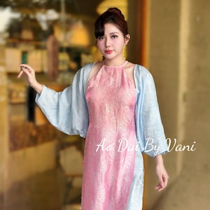 May include: A pink halter-style dress with a floral pattern, paired with a light blue shawl. The dress has a high neckline and a long, flowing design. The text "Ao Dai By Vani" is visible on the dress.