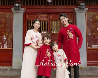 May include: A family of four wearing traditional Vietnamese clothing, known as Ao Dai, in red and cream colors. The clothing features intricate embroidery and the family is standing in front of a red building with a gold trim.
