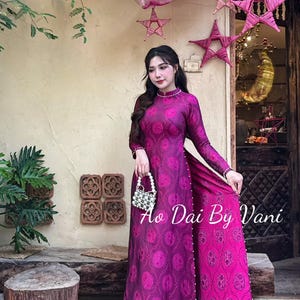 May include: A magenta Ao Dai dress with a circular pattern and long sleeves. The dress has a high neckline and is adorned with small pearls. The text "Ao Dai By Vani" is visible on the dress. The dress is worn with sandals.