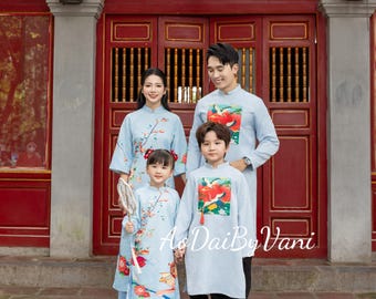 May include: A family of four wearing matching light blue traditional Vietnamese Ao Dai outfits. The outfits feature floral and bird designs. The text "Ao Dai By Vani" is visible. The background is a red and brown building.