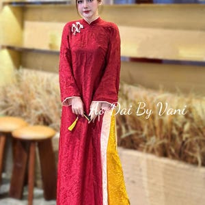 May include: A red Ao Dai dress with long sleeves and a mandarin collar. The dress has a textured pattern and white trim on the cuffs. The lower portion of the dress is yellow with a white stripe. The text "Ao Dai By Vani" is visible.