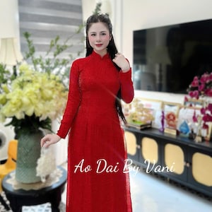 May include: A long, red dress with long sleeves and a high neckline. The dress has a textured pattern and the text "Ao Dai By Vani" is visible. The woman is standing in front of a blurred background.