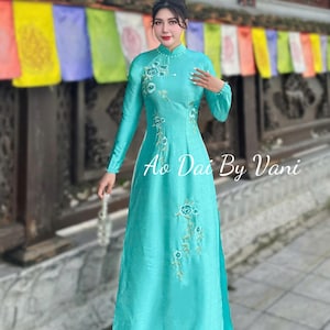 May include: A turquoise Ao Dai dress with long sleeves and floral embroidery. The dress features a high neckline and a flowing skirt. The text "Ao Dai By Vani" is visible on the dress. The dress is made of a silky material.