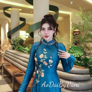 May include: A teal-colored Ao Dai dress with long sleeves and floral embroidery. The dress features a beaded collar and the text "AoDaiByVani" in white script. The dress has an orange lining and is made of a shiny fabric.
