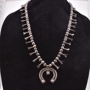 Puede incluir: Un collar de plata de flor de calabaza exhibido en un maniquí negro. El collar presenta una serie de cuentas redondas y colgantes de flores estilizadas. El colgante tiene forma de media luna con detalles decorativos.