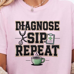 May include: A light pink t-shirt with the words "DIAGNOSE SIP REPEAT" in a black and gold design. A stethoscope, clipboard, and coffee cup are also featured in the design.