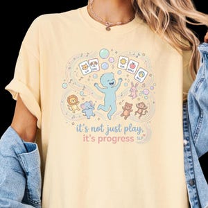 Könnte beinhalten: Hellgelbes T-Shirt mit farbenfroher Grafik. Das Design zeigt eine blaue Figur, Cartoon-Tiere und Objekte. Der Text auf dem Shirt lautet "it's not just play, it's progress SLP."