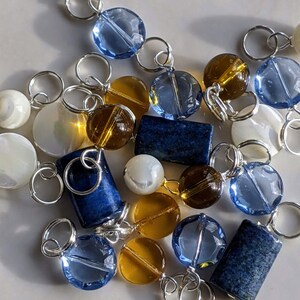 May include: A collection of blue, brown, and white beads and charms, including round and rectangular shapes, strung on silver jump rings.