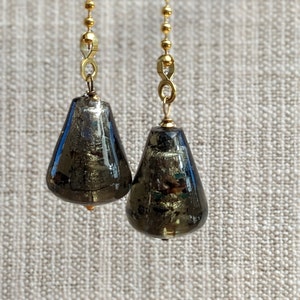 May include: Two decorative ceiling fan pull chains. Each chain has a teardrop-shaped, dark gray glass pull with gold accents, suspended from a gold beaded chain. The background is a textured, neutral fabric.