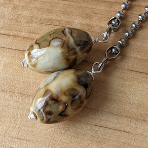 May include: Two oval-shaped, marbled beads in shades of brown, tan, and cream, suspended from a silver-colored chain. The beads have a glossy finish and are connected to the chain with silver-toned metal findings. The background is a light brown wooden surface.