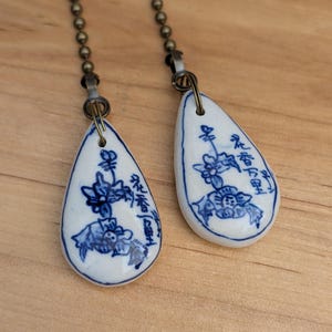 May include: Two teardrop-shaped, white ceramic light pulls with blue floral designs and Asian characters. Each pull is attached to a bronze-colored ball chain. The background is a light brown wooden surface.