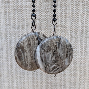 May include: Two round, wooden-look ceiling fan pull chains with black beaded chains. The circular pulls have a marbled, gray and brown pattern. The background is a neutral, textured fabric.