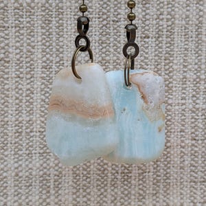 May include: Two ceiling fan pulls featuring irregular, polished stones with bands of pale blue, white, and tan. Each stone is suspended from a bronze-toned metal jump ring attached to a matching bronze-toned ball chain connector and chain.