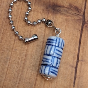 May include: A blue and white ceramic bead with a geometric pattern hangs from a silver chain with a silver clasp.