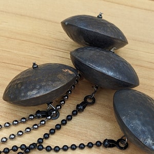 May include: Three dark, hammered metal dome-shaped ornaments, each attached to a thin ball chain. Two chains are black, and one chain is a metallic gunmetal gray. The domes have a textured, uneven surface with a dark, weathered finish.
