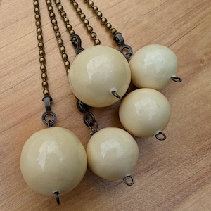 May include: A set of five cream-colored, spherical ceramic beads attached to individual antique brass-toned ball chains. Each bead features a glossy finish and a small metal eyelet at the bottom, suspended from the metallic chains in a cluster arrangement.