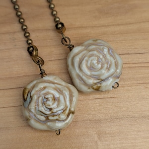 May include: Close-up of a necklace featuring two cream-colored rose pendants. The pendants have a glossy finish with brown accents and are suspended from a bronze-colored chain. The background is a light brown wooden surface.